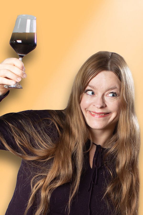 Mischievous woman with long hair holding a stemmed glass with dark liquid.