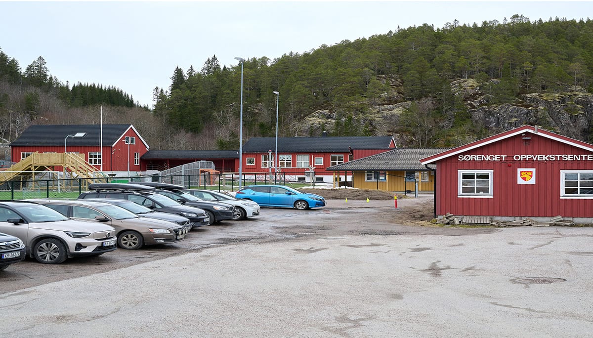 Red buildings of SØRENGET OPPVEKSTSENTER, a parking lot with various cars, and a wooded hillside.