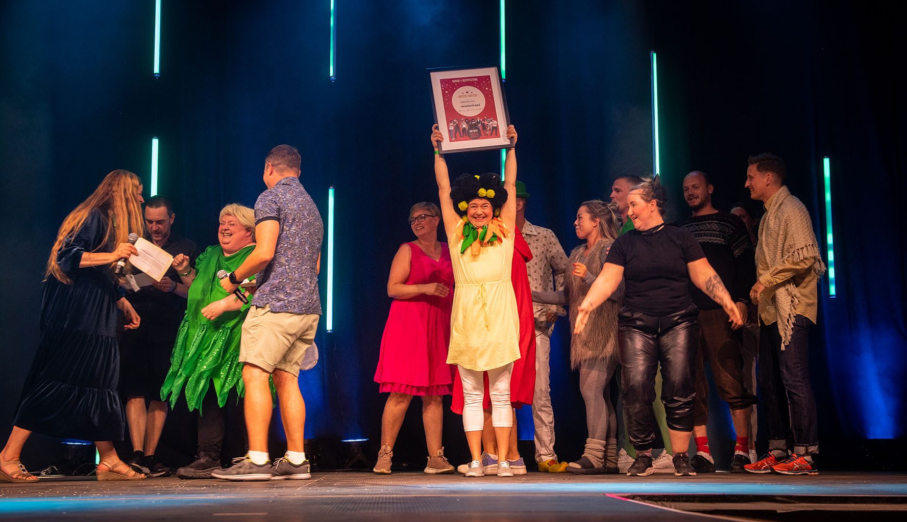 A diverse group of people on a stage celebrating, one person holding a framed award over their head.
