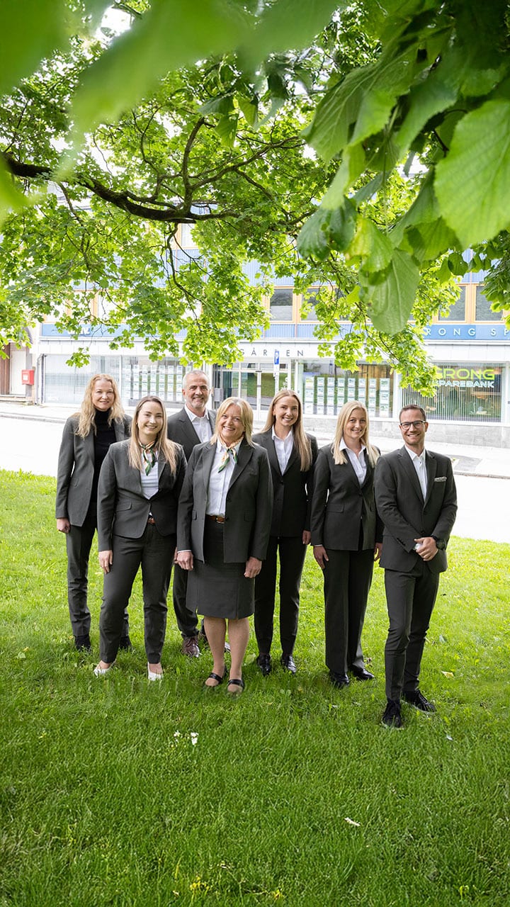 A smiling group of seven people in dark suits standing on green grass outdoors.