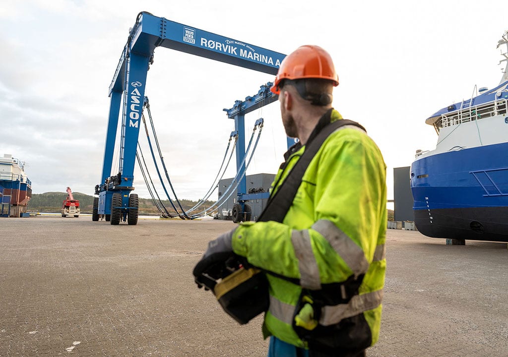 Worker in hard hat and hi-vis vest operates remote for a large blue gantry crane at a marina.