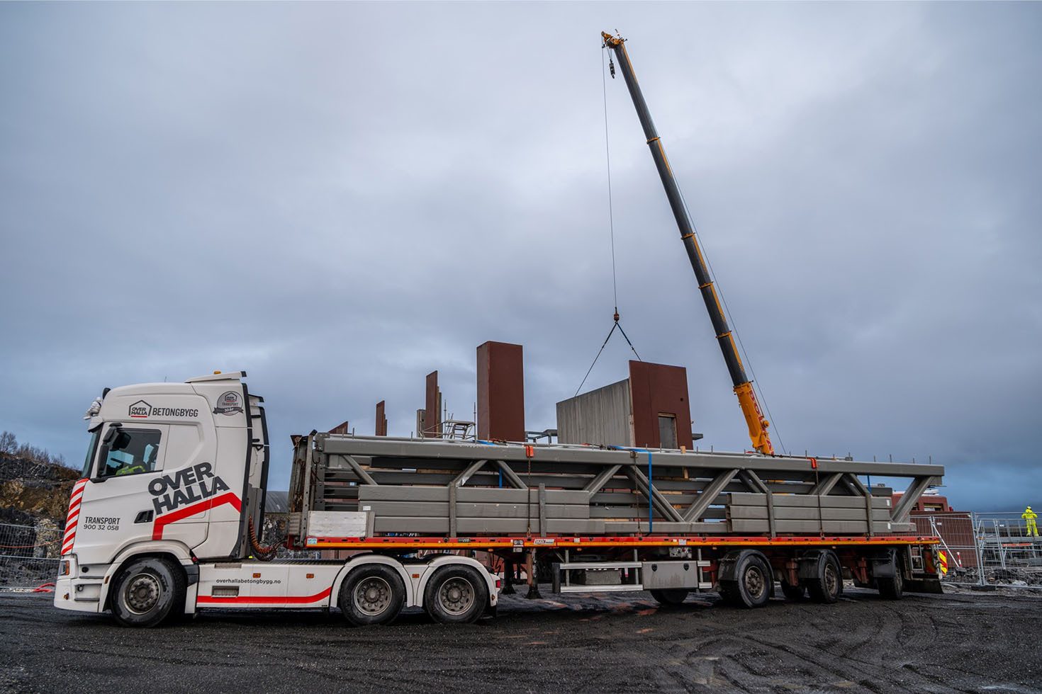 Crane lifting a concrete panel on a construction site next to a truck carrying metal trusses.