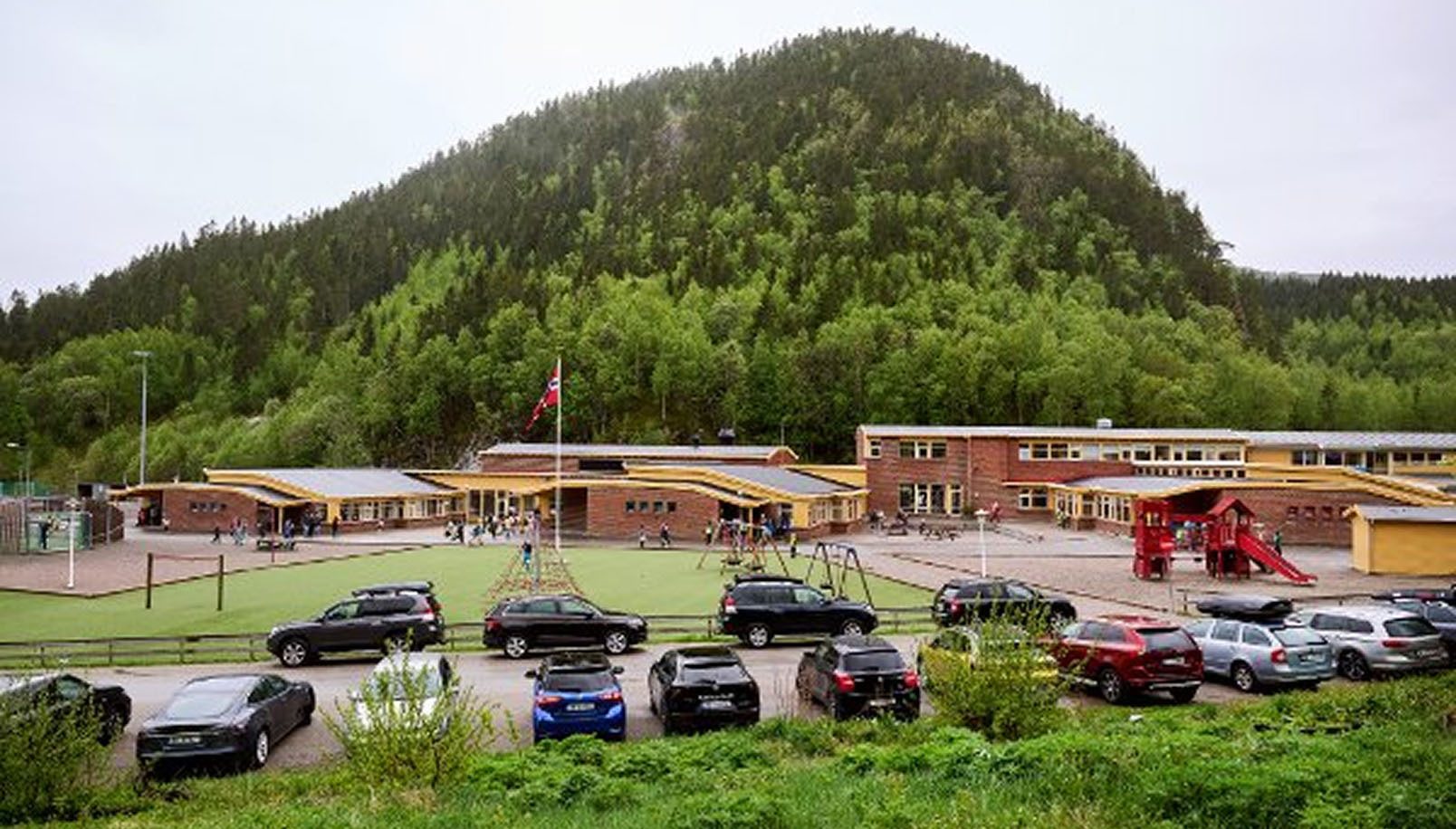 A school building and playground with children, a parking lot, and a large forested hill in the background, under an overcast sky.