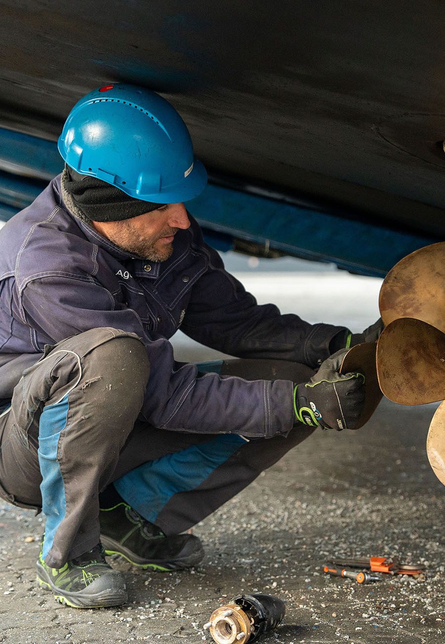 Man in blue hard hat and gloves inspects boat propeller under hull.