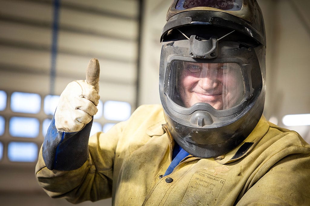Welder smiles through helmet, gives thumbs-up.