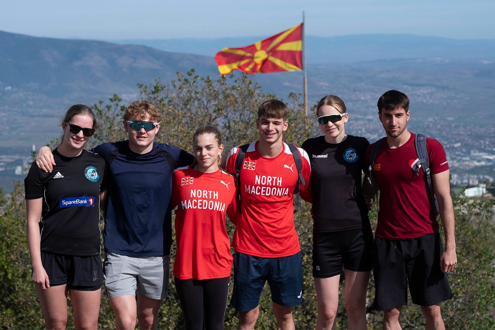 Six athletes in North Macedonia gear pose on a mountain, with the national flag and cityscape visible.