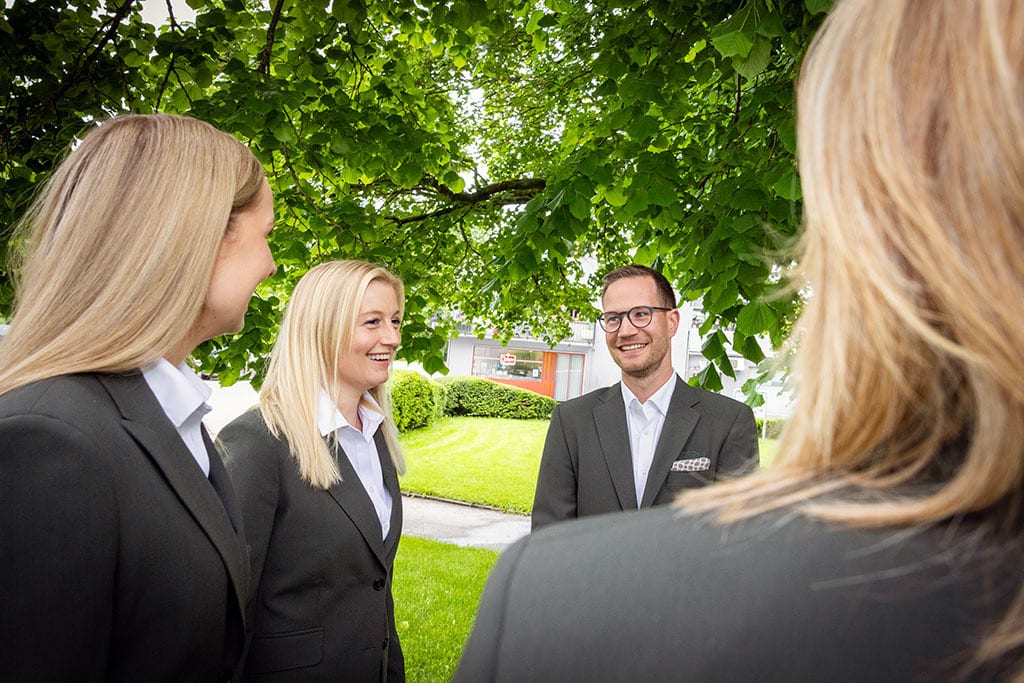 Four smiling business people conversing outdoors under trees.