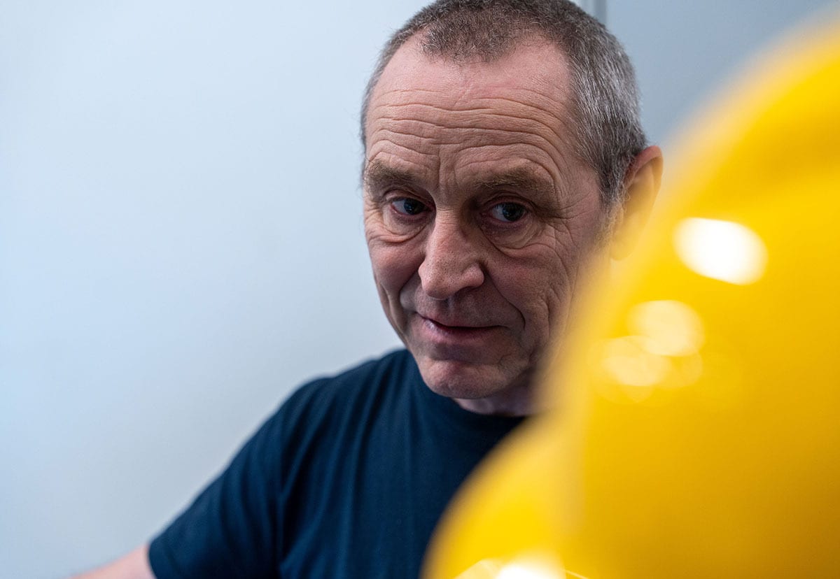 A man with grey hair and wrinkles in a dark shirt looks at the camera, blurred yellow on right.