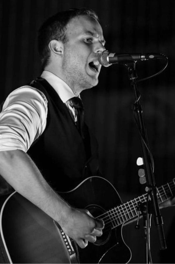 Black and white photo of a man singing into a mic while playing an acoustic guitar.