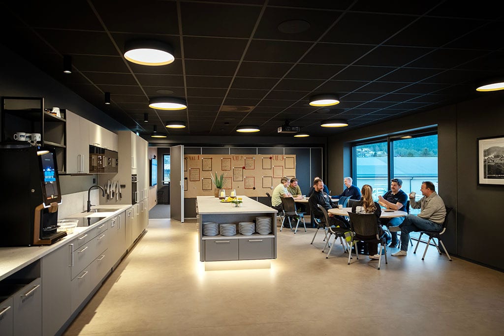 Modern office breakroom with people socializing, a kitchen, and scenic window view.