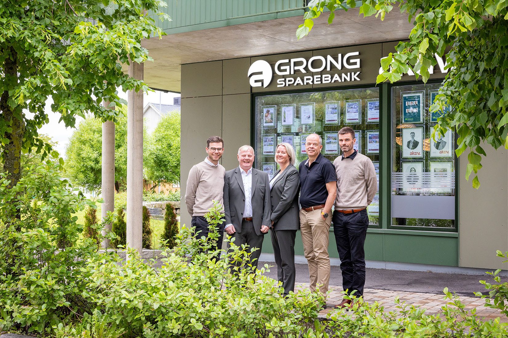 Five people (3 men, 2 women) stand in front of the Grong Sparebank building with greenery.