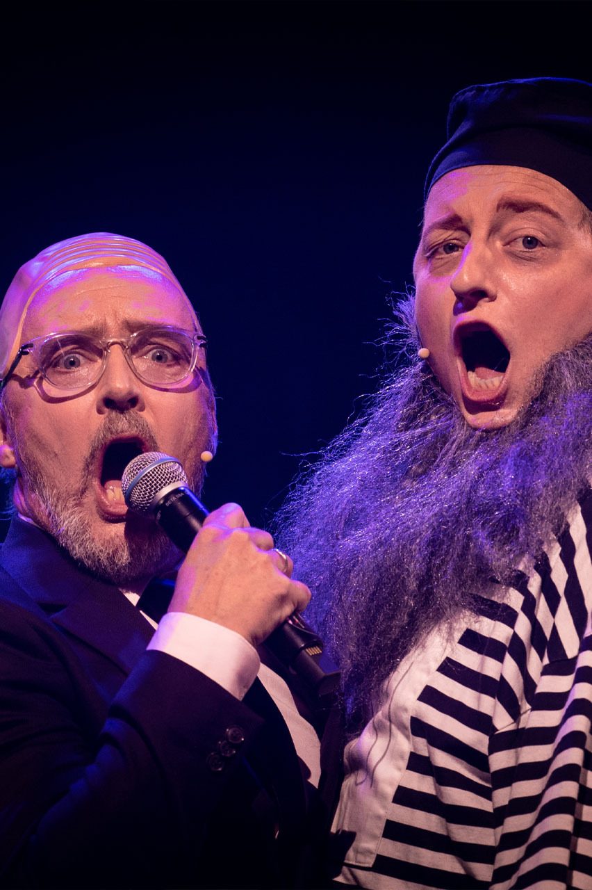 Two theatrical performers, mouths open, singing. One bald man in a suit, the other with a grey beard and stripes.