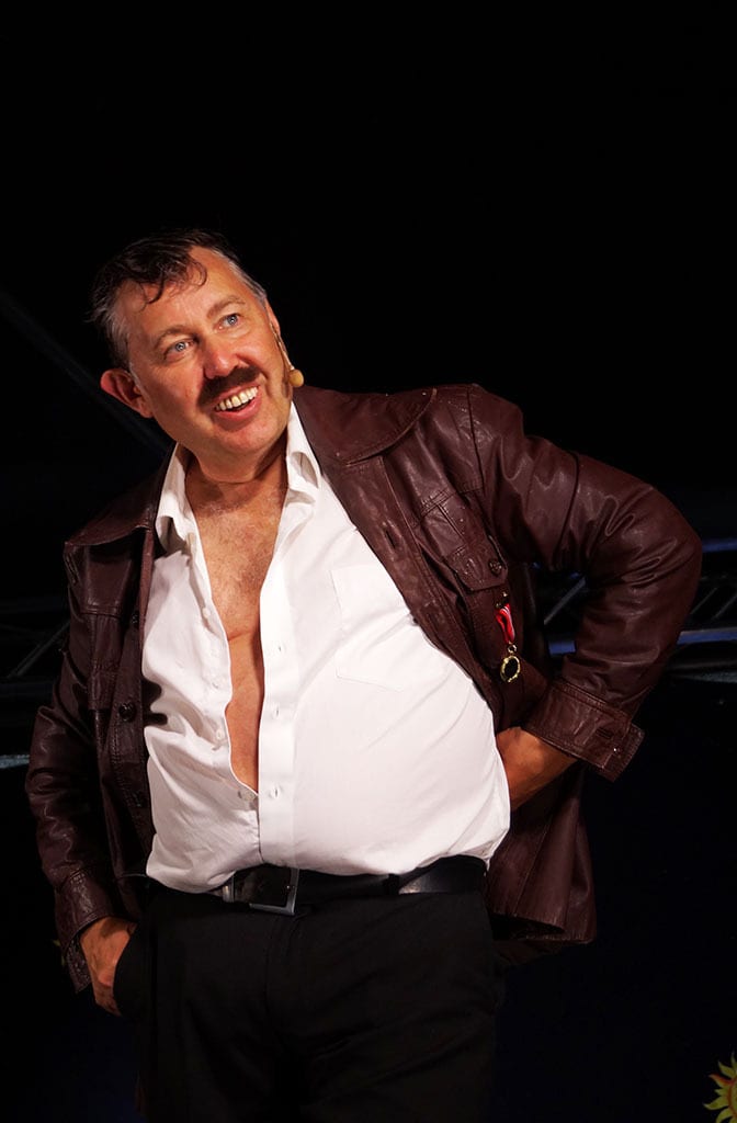 Smiling man with mustache in brown jacket and open white shirt looking up.