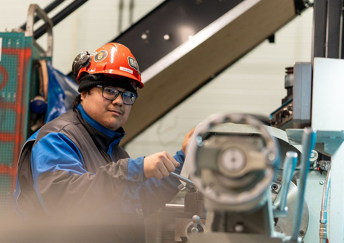 Man in an orange hard hat and glasses operating industrial machinery, looking at the viewer.