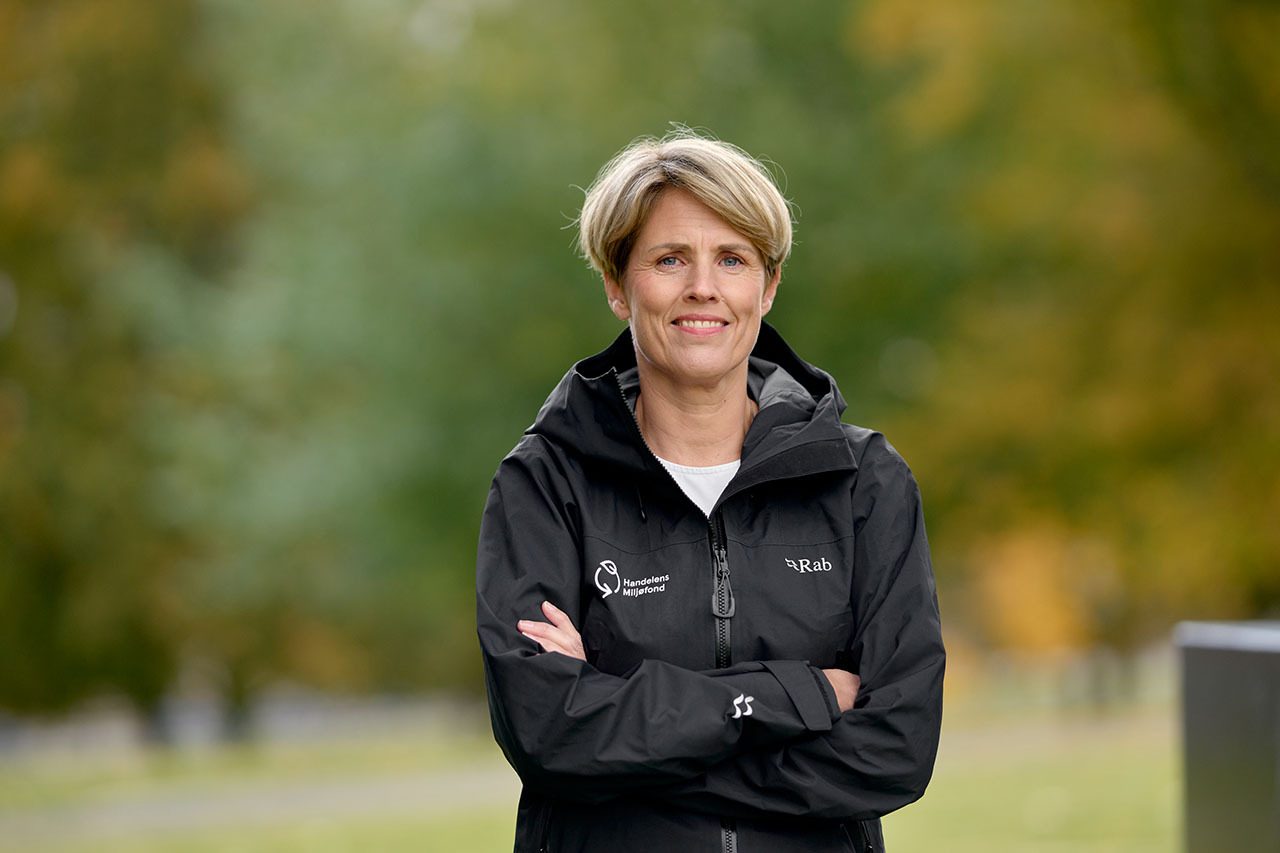 Smiling woman with short blonde hair, wearing a black jacket, arms crossed, outdoors.