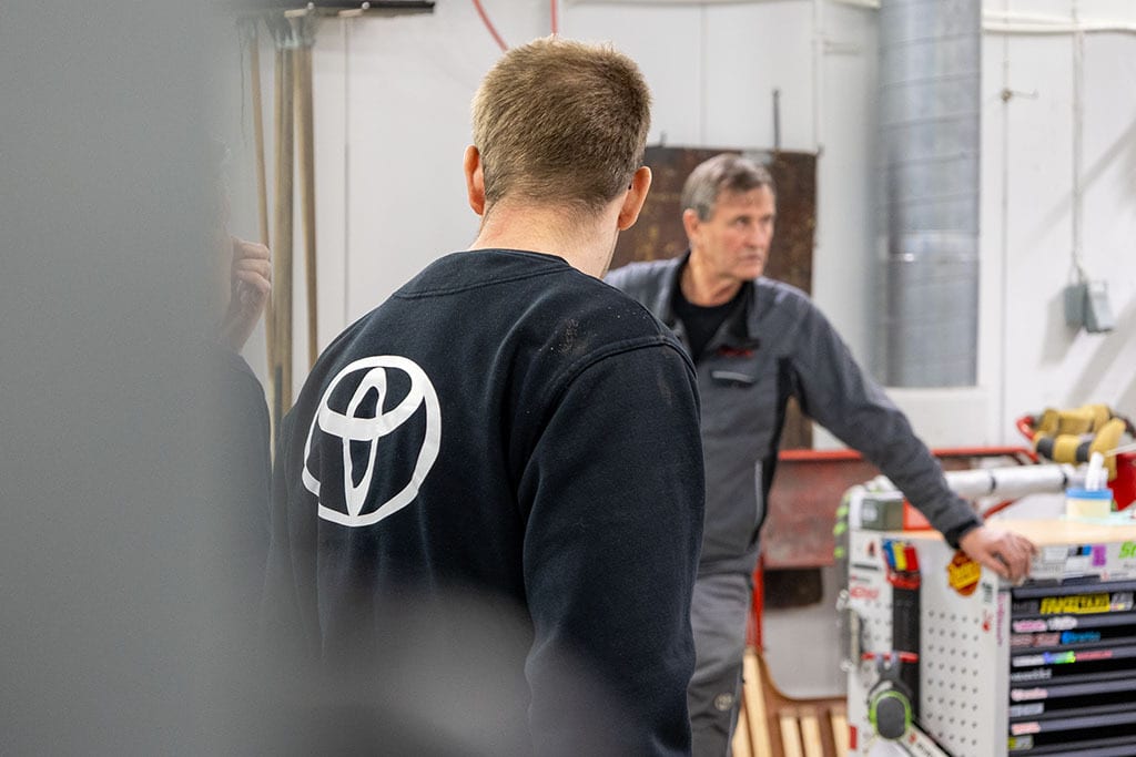 Back of a person wearing a black Toyota sweatshirt, another man in a workshop.