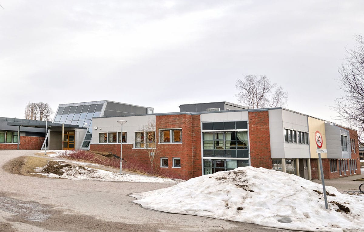 Modern school building with brick and grey facades, angular glass sections, snow piles, and bare trees under a cloudy sky.