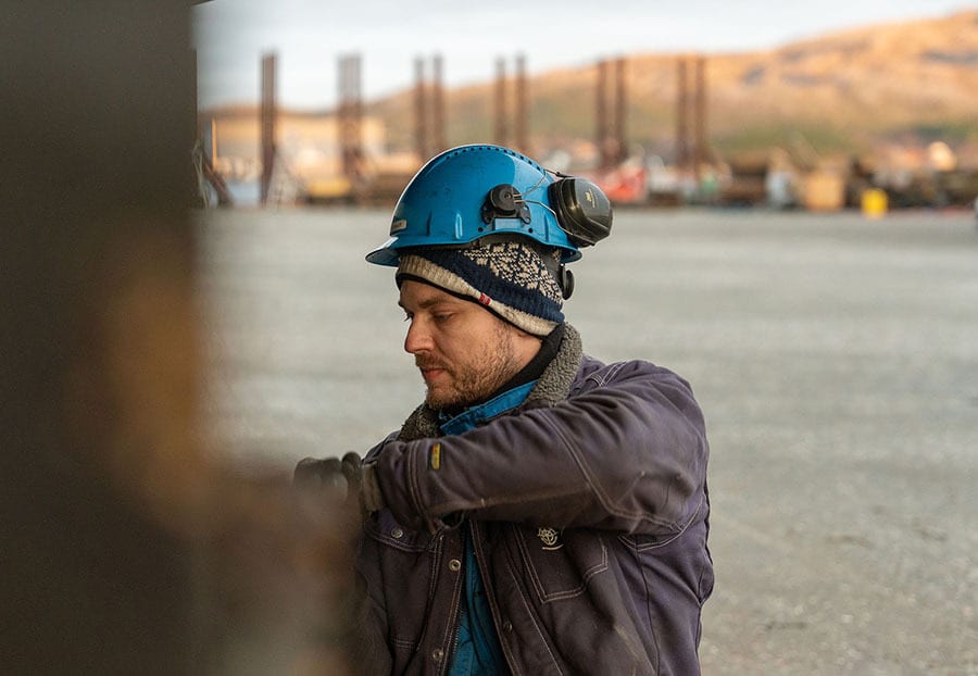 Worker in blue hard hat and beanie, looking down, outdoors at an industrial site.