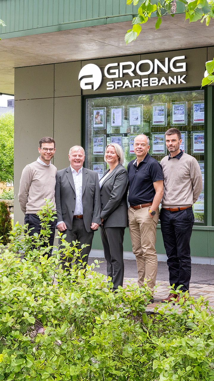 Five people in front of Grong Sparebank, with bushes in the foreground.