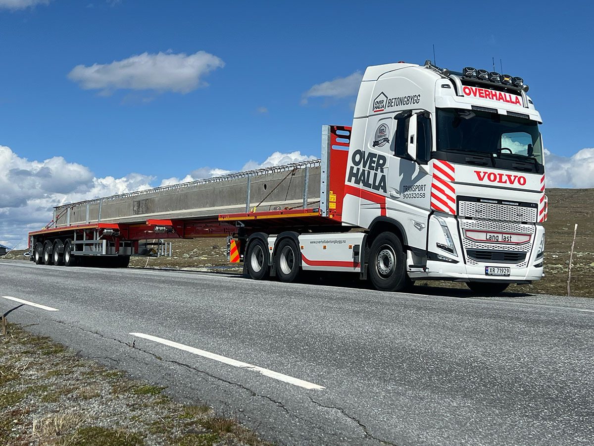 Over Halla Volvo truck with a long concrete beam on a flatbed trailer on a road.