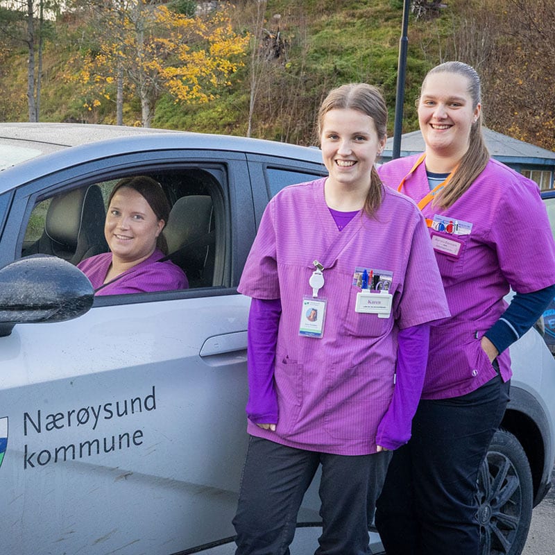 Three smiling women in purple scrubs, two standing, one in a "Nærøysund kommune" car.