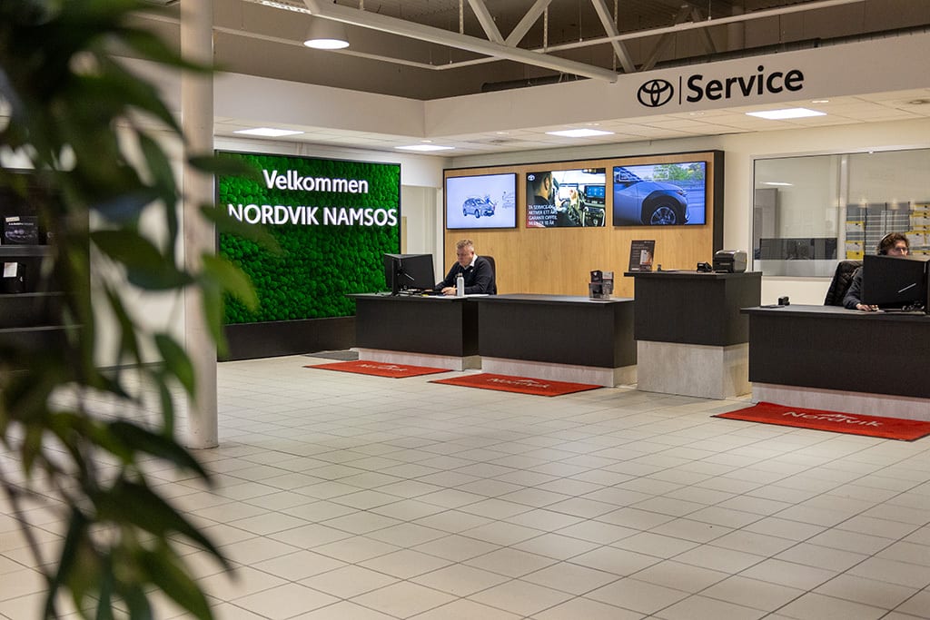 Toyota service reception with two employees, a moss "Velkommen" sign, and digital car displays.