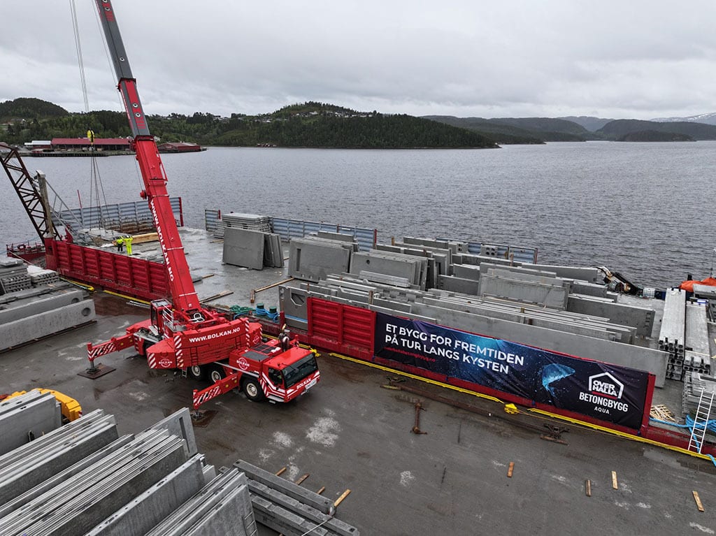 Red mobile crane loading concrete slabs onto a barge at a dock by the water, with a banner.