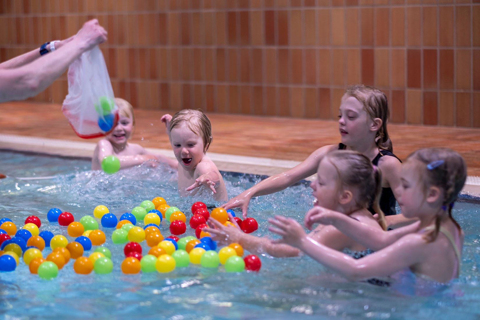 Children play excitedly with colorful balls in a swimming pool, while an adult adds more from a bag.