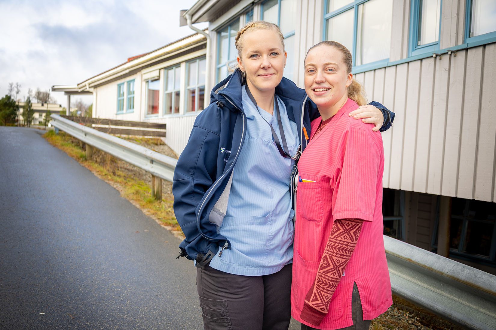 Two smiling women in medical attire stand outdoors, one with her arm around the other.