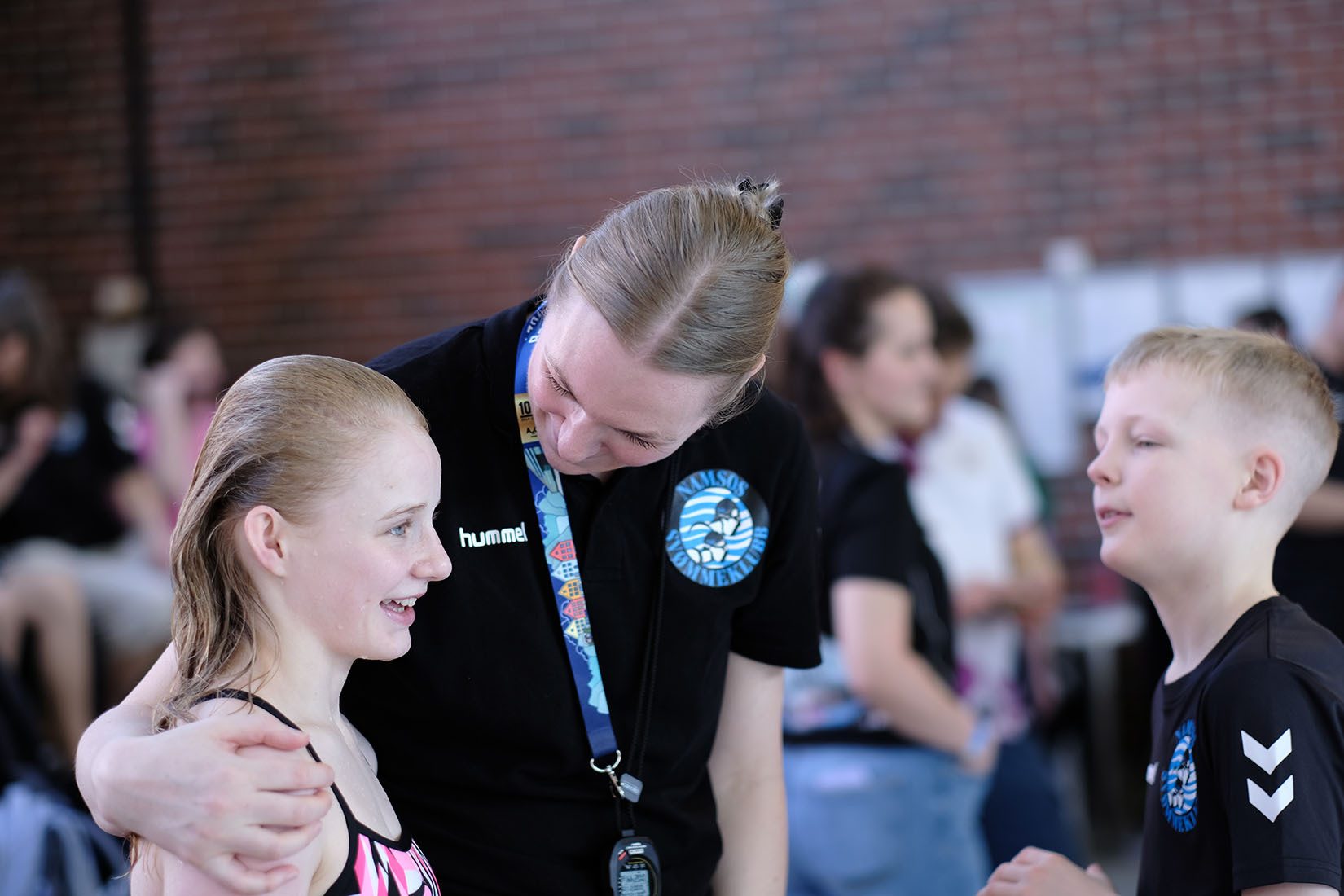 A swimming coach interacts with two young swimmers, a girl and a boy, after a swim.