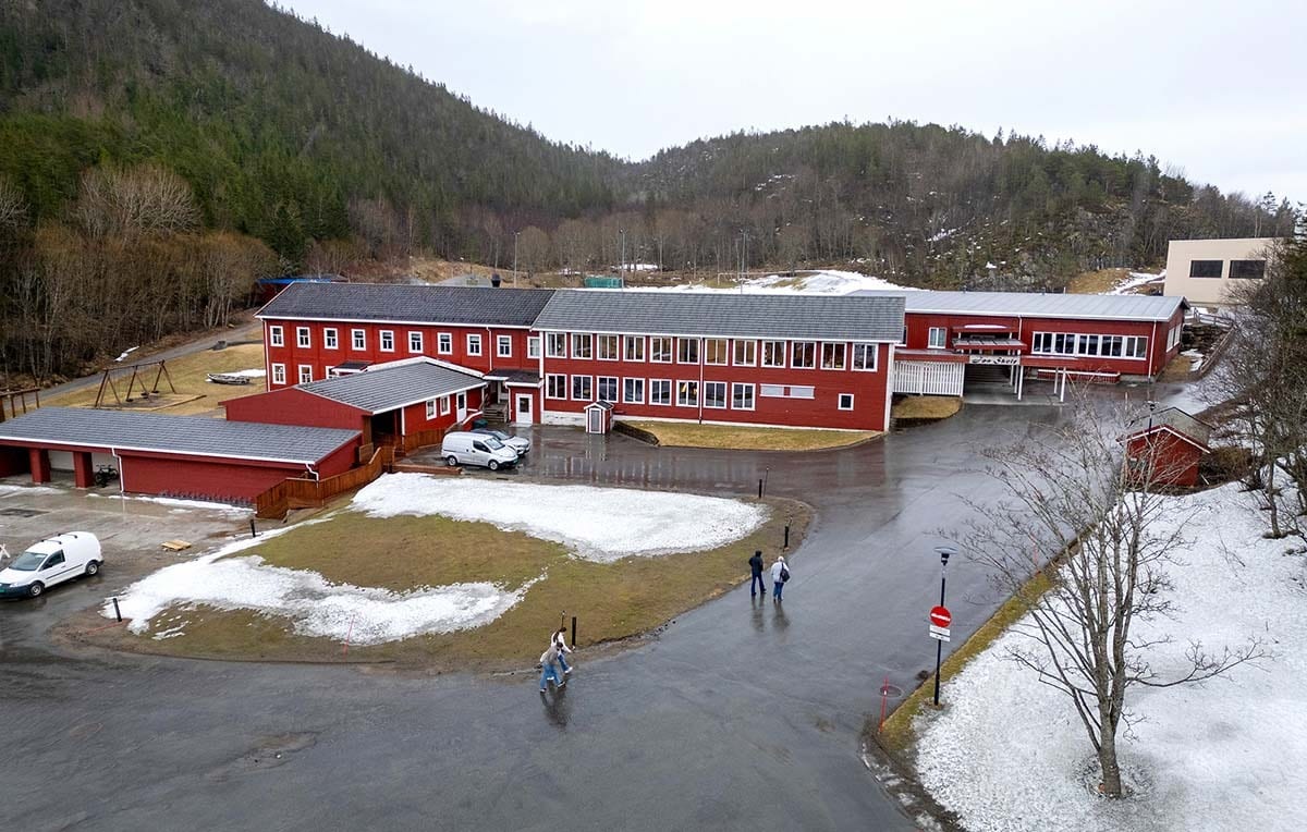Large red building complex with multiple sections and white vans on a partially snow-covered ground with a forested mountain backdrop.