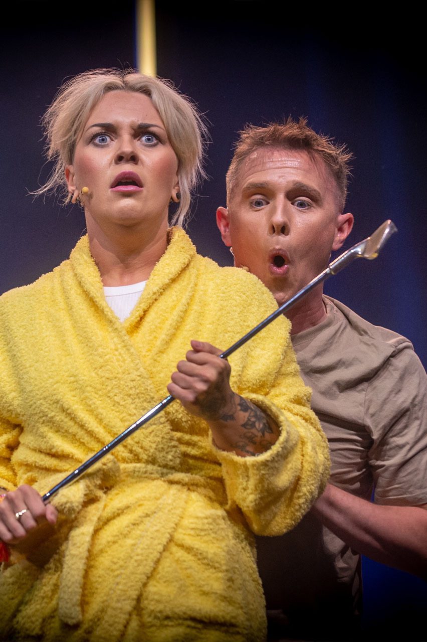 Woman in yellow bathrobe and man behind her look comically shocked. She holds a golf club.