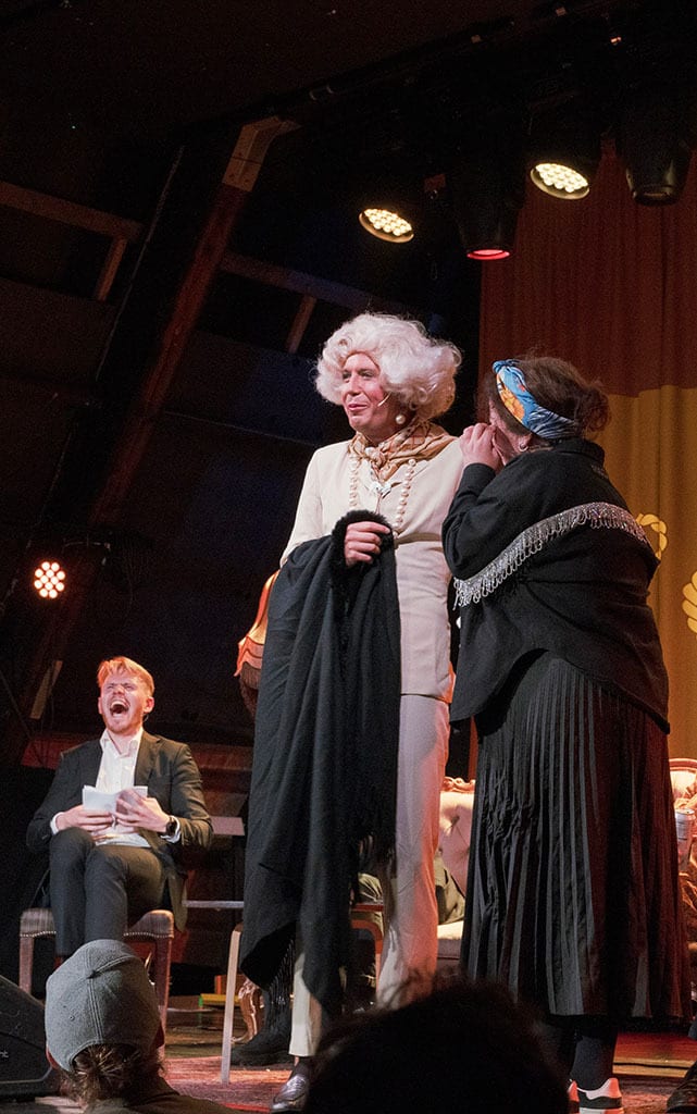 White-wigged performer on stage, whispered to by another, as a man laughs.