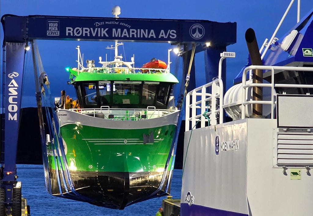A green boat is lifted by a blue Rørvik Marina gantry crane, next to a white boat.