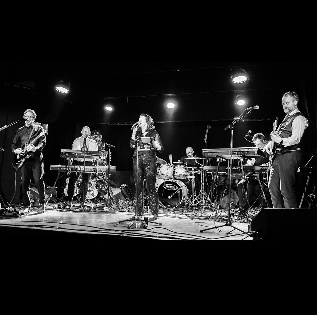 Black and white band: female singer, 2 guitarists, 2 keyboardists, and a drummer on stage.