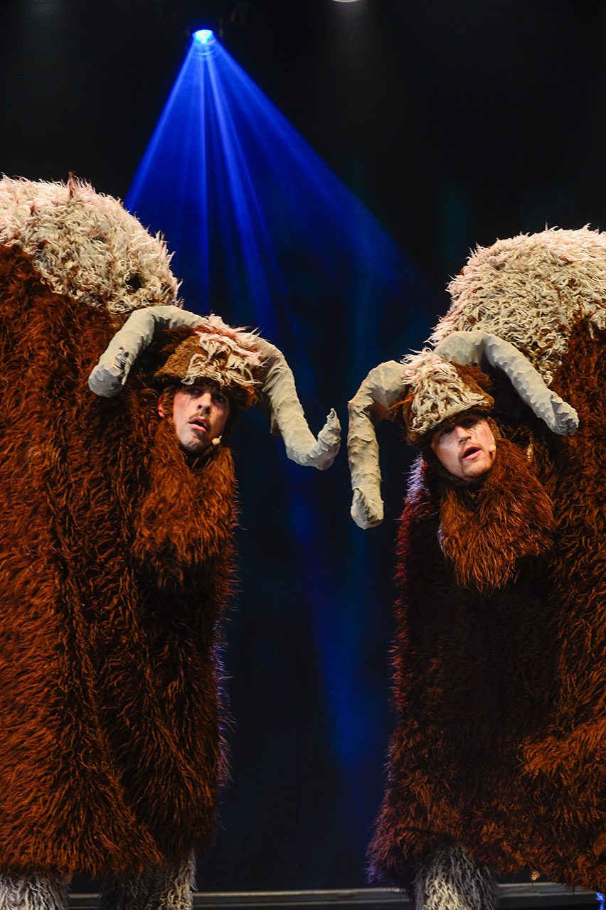 Two performers in brown, furry musk ox costumes with horns under a blue spotlight.