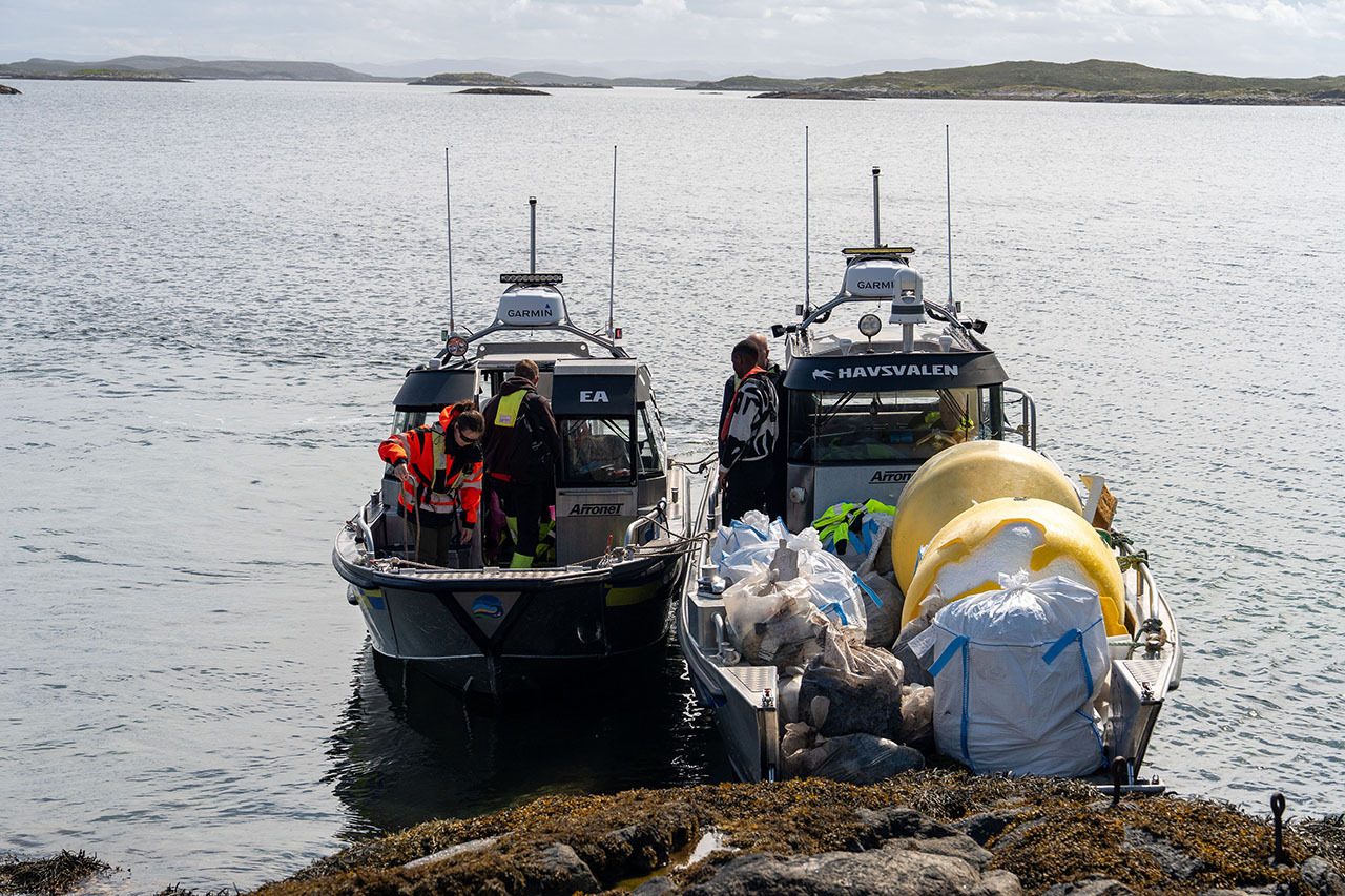 Two boats with people and collected marine debris docked at a rocky shore.