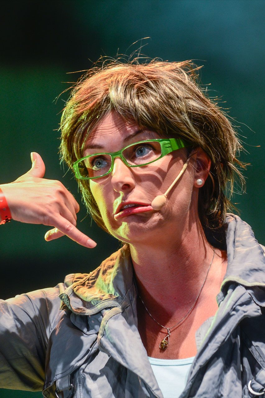 Woman in green glasses and headset mic makes "call me" gesture with comical expression.