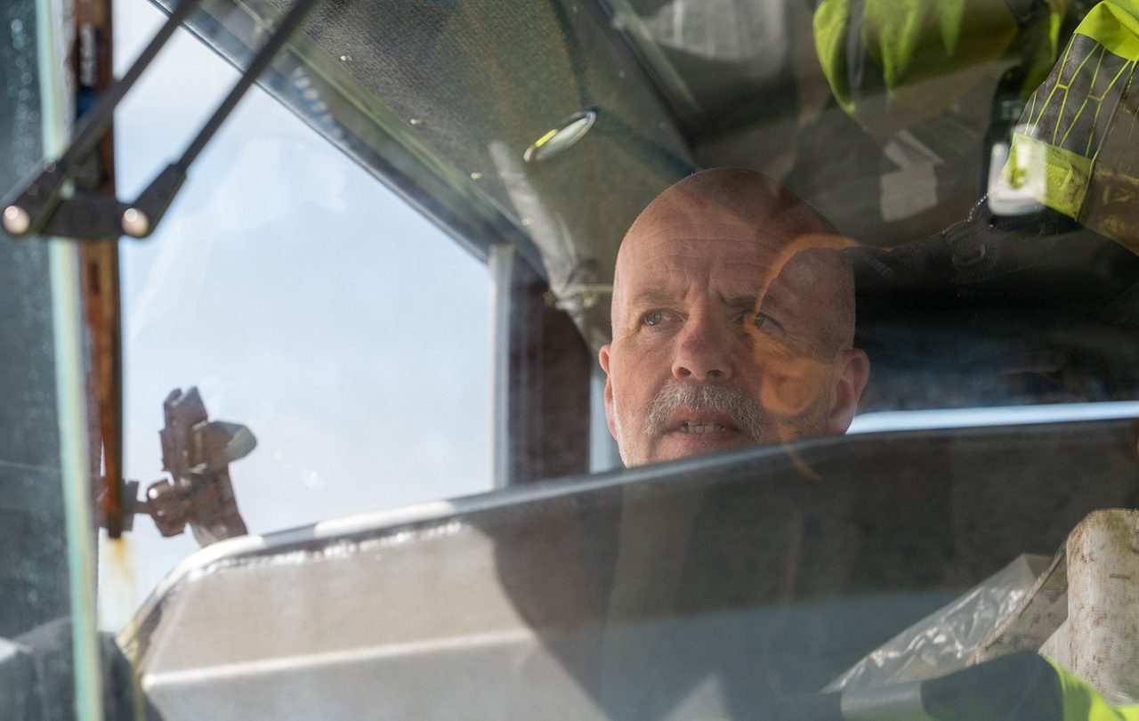 Bald man with mustache looking intently through a window, reflections on glass.