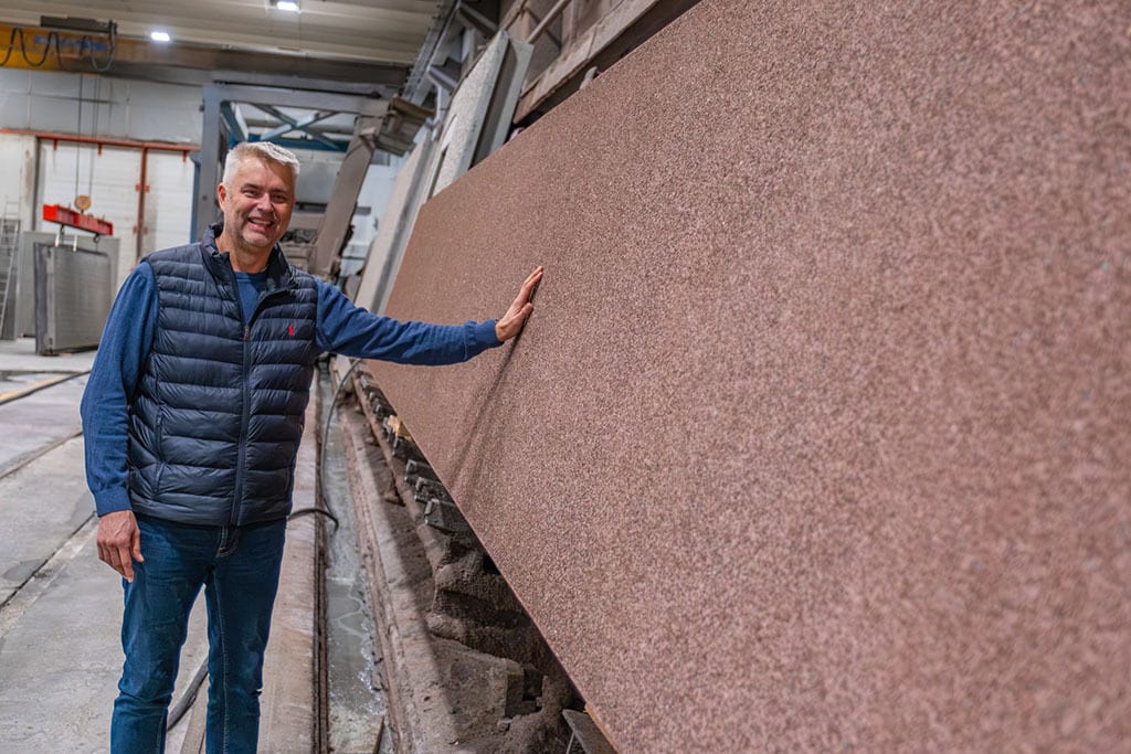Smiling man in a factory touches a large, reddish stone slab.