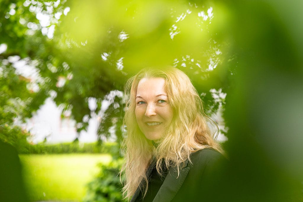 Smiling blonde woman framed by green leaves outdoors.