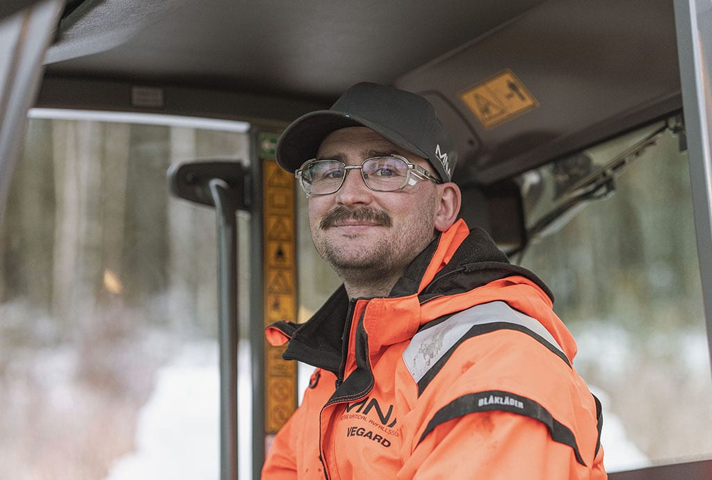 Smiling man with mustache, wearing glasses, cap, and orange work jacket, inside a vehicle.