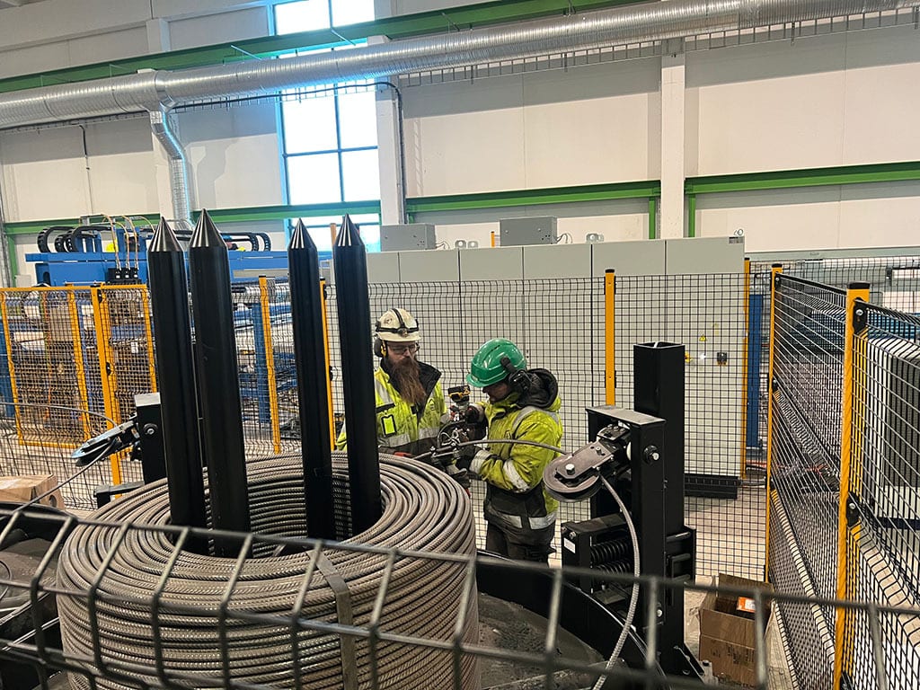 Two men in safety gear work with rebar coils and industrial machinery in a factory.