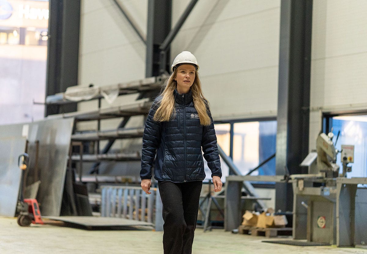 Woman in hard hat and puffer jacket walks in a factory.