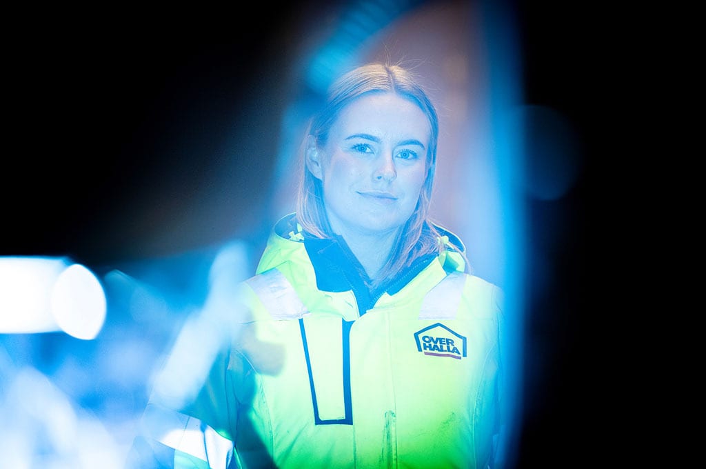 Young woman in yellow high-vis jacket with 'OVER HALLA' logo, framed by blue light effects.