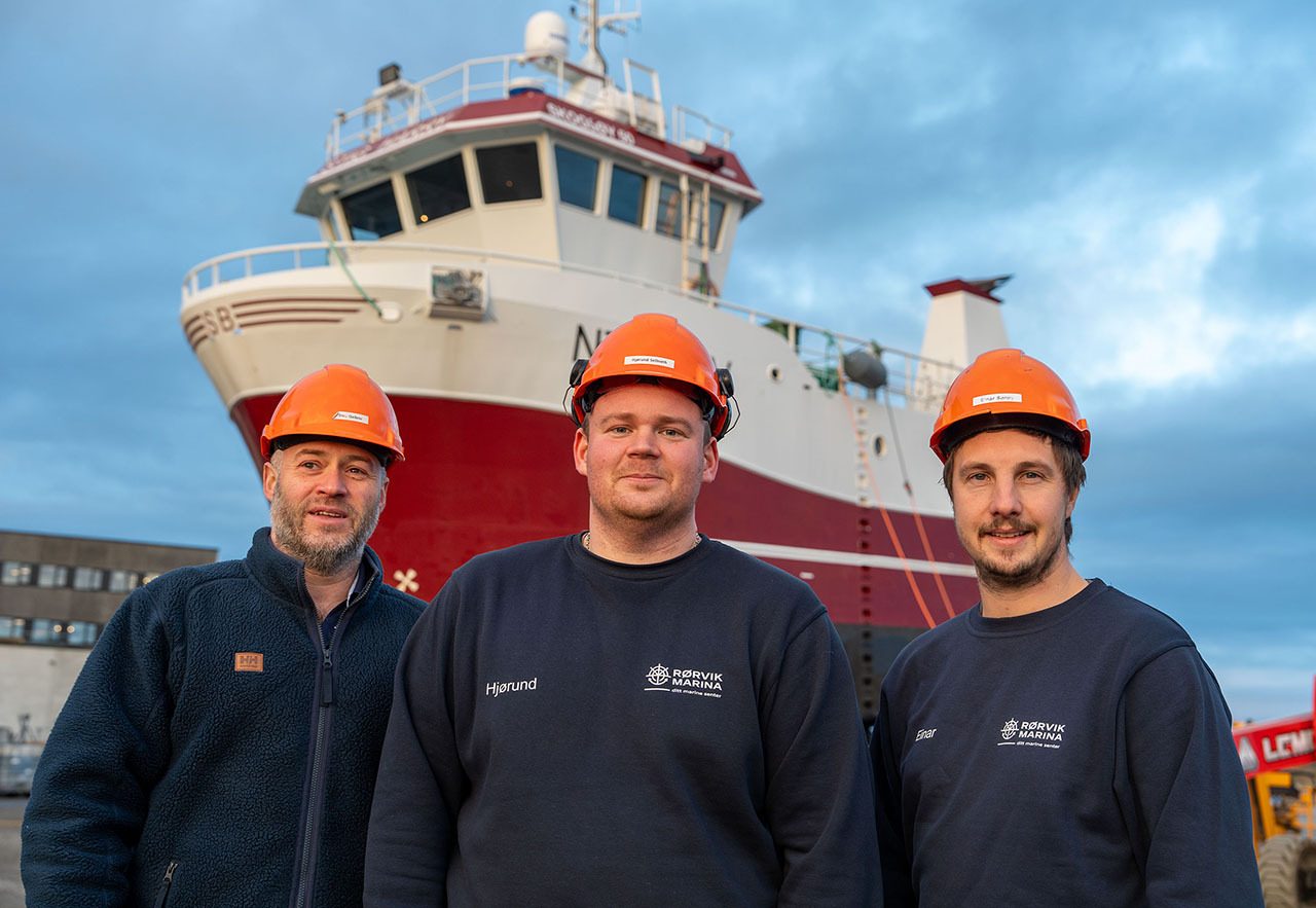 Three men in orange hard hats and dark shirts standing in front of a red and white boat.
