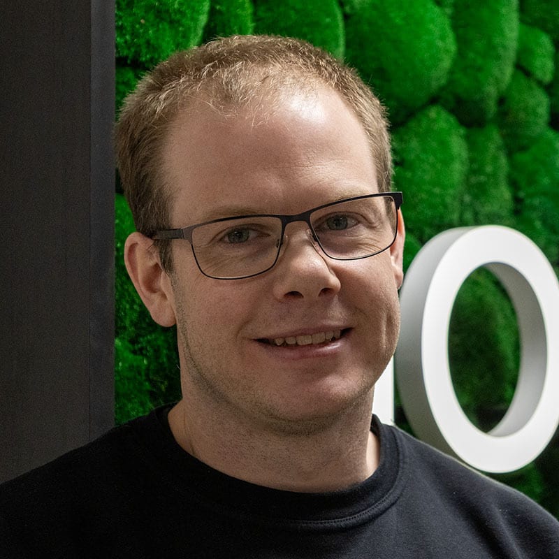 Smiling man with glasses, short light hair, black shirt, green textured background with white letter 'O'.