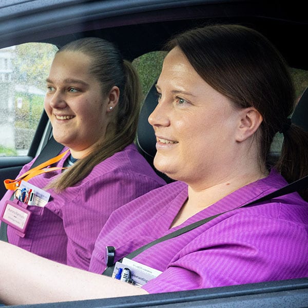 Two smiling healthcare professionals in purple scrubs in a car.