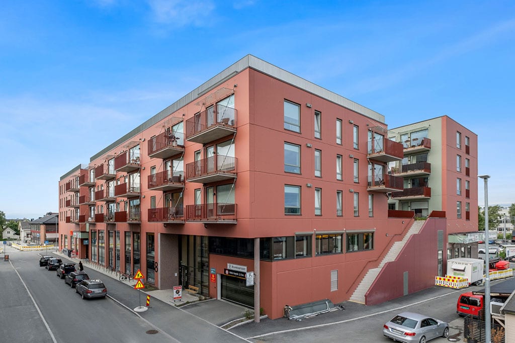 Modern, red-orange apartment building with balconies, many windows, and a street with parked cars.