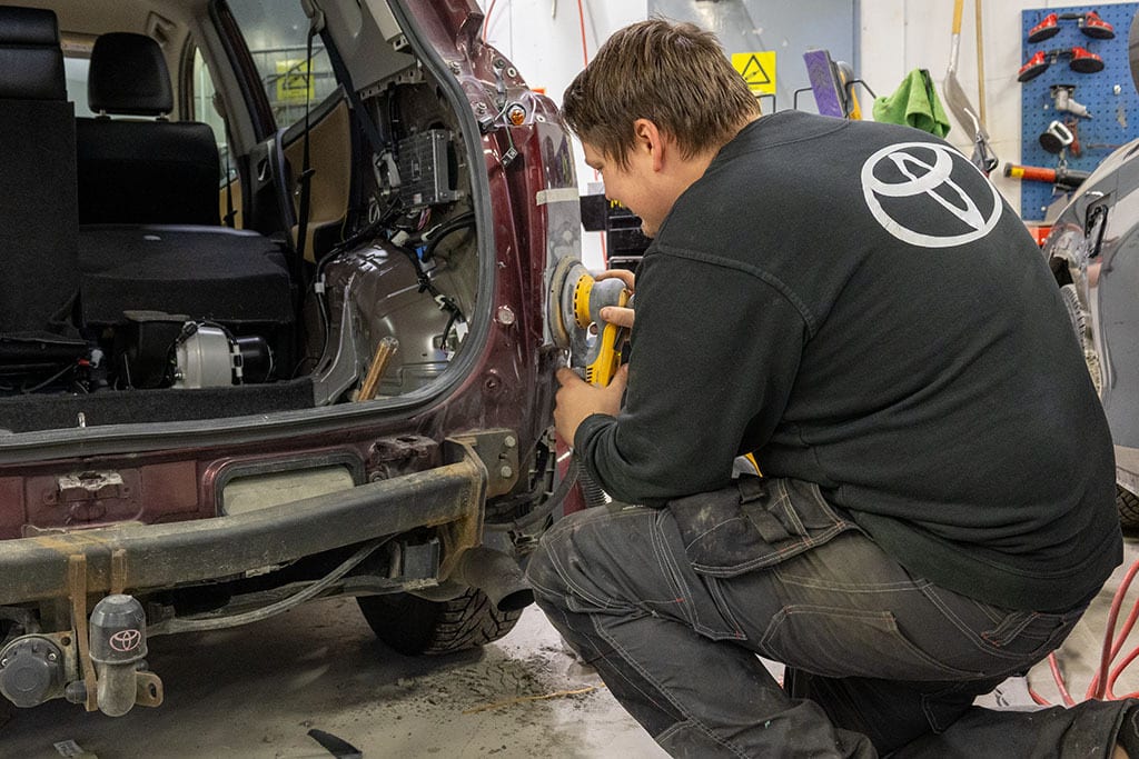 Man in a Toyota shirt sanding a car's rear quarter panel in a garage.